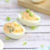 Kimchi spicy deviled eggs lined up with green onions on top and a bowl of kimchi behind them with more eggs on a cutting board.