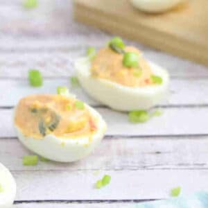Kimchi spicy deviled eggs lined up with green onions on top and a bowl of kimchi behind them with more eggs on a cutting board.