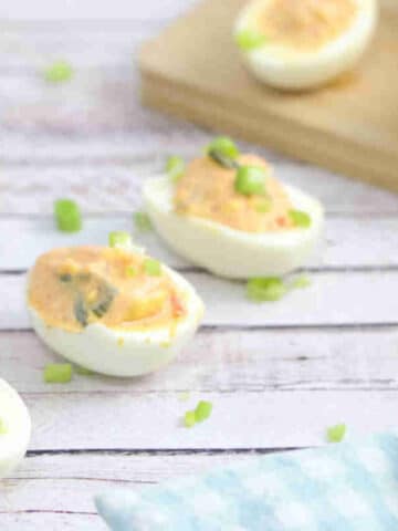 Kimchi spicy deviled eggs lined up with green onions on top and a bowl of kimchi behind them with more eggs on a cutting board.