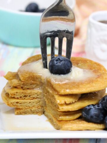 five banana-free protein pancakes stacked with blueberries beside them and butter on top with a fork stuck in them.