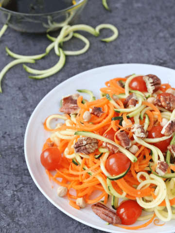 Spiralized zucchini and carrotes with avocado, cherry tomatoes and strawberries on a white plate with dressing on the side