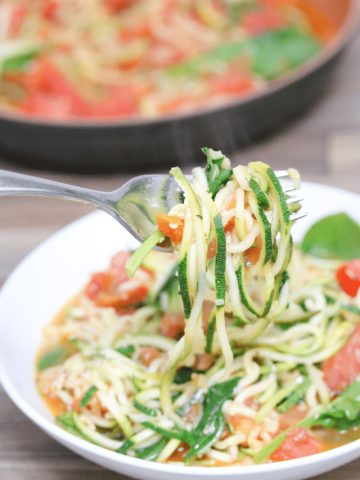 White bowl with a fork of zucchini noodle pasta and tomatoes.