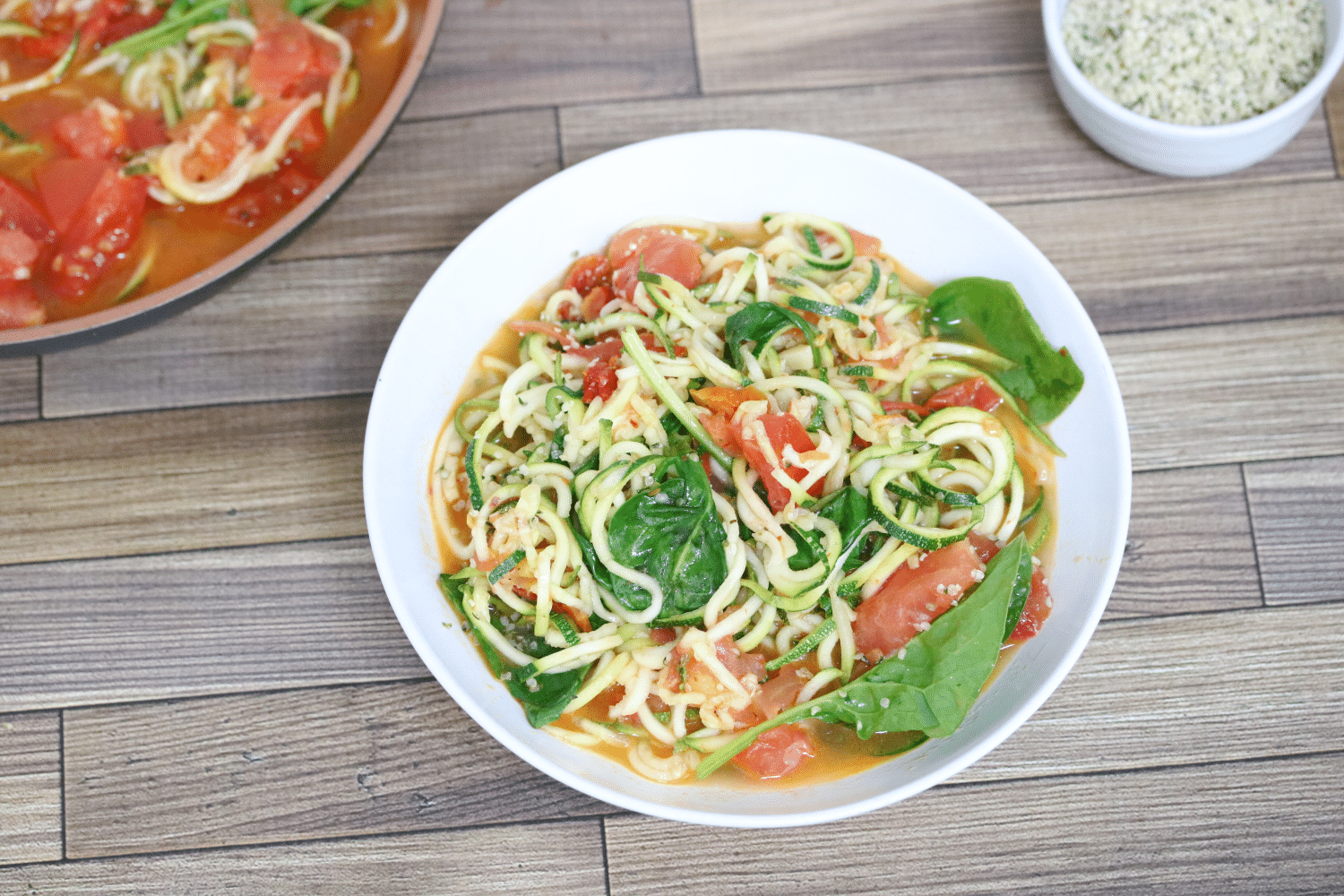 Zucchini and tomato pasta with spinach in a white bowl with more in a pan behind it