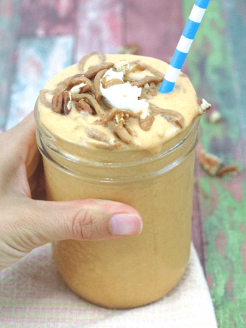 Pin image with text: orange pumpkin pie smoothie in a mason jar topped with caramel sauce, coconut cream and a blue straw being held in a hdn