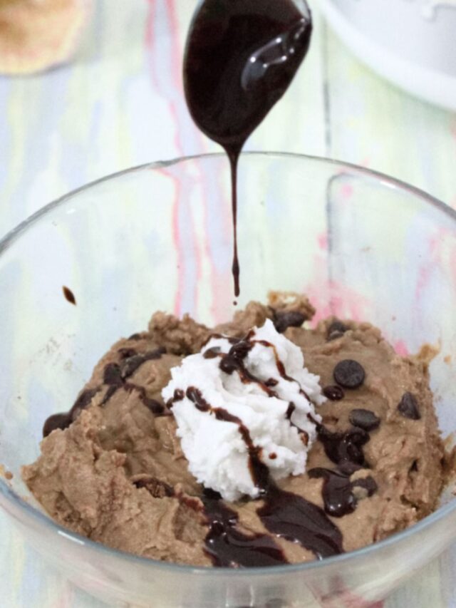 Spoon dripping chocolate onto a glass bowl of chocolate protein fluff topped with coconut whipped cream.