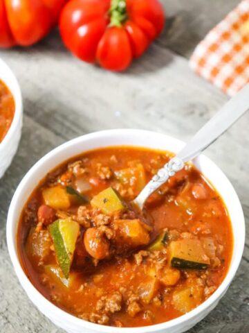 white bowl with venison and zucchini chili and a spoon in the bowl with ingredients behind it