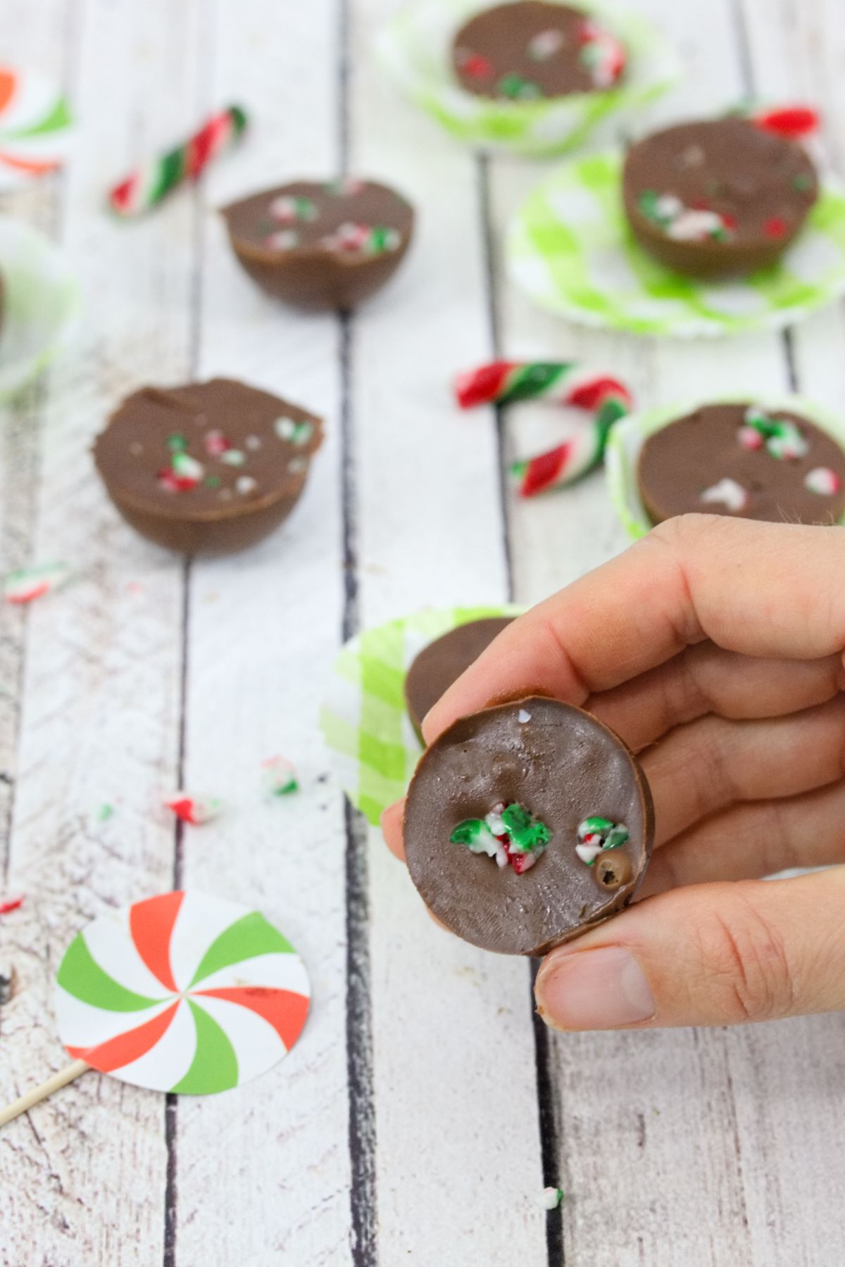 Hand holding small portion of chocolate fudge with candy canes on top with more fudhe on green cupcake holders behind it.