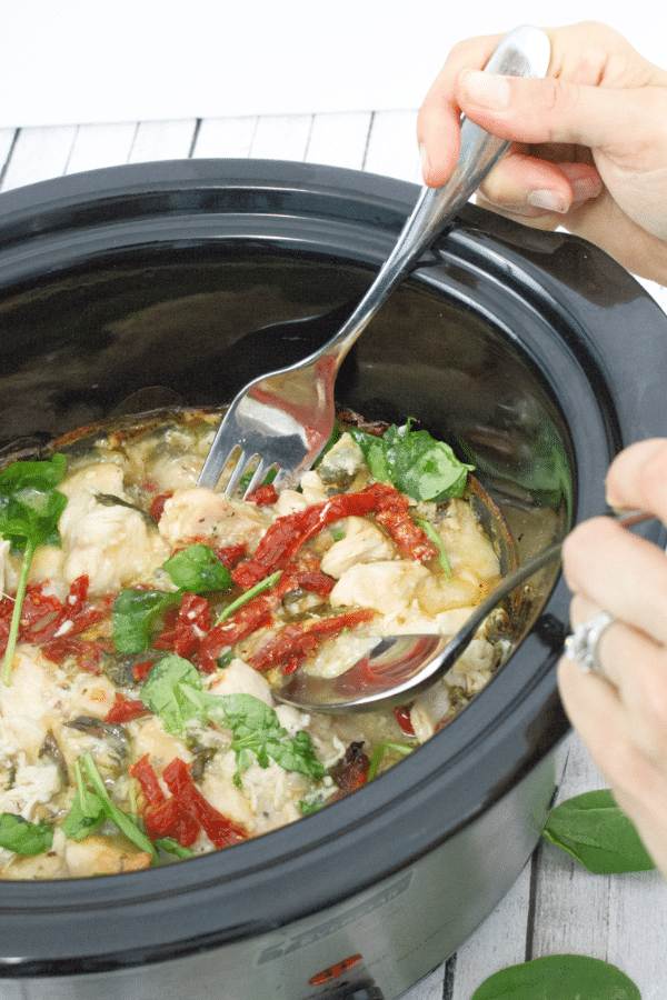 Pin image with text: creamy tuscan chicken in a bl;ack crockpit with sundried tomatoes, basil and a spoon and fork in the slow cooker