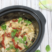 Pin image with text: black slow cooker with dairy-free tuscan chicken, sause, sundried tomatoes and basil with the lid on a colored linen beside it