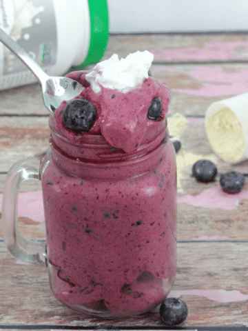 Mason jar with puepl protein fluff topped with blueberries and coconut cream and a spoon scooping into it