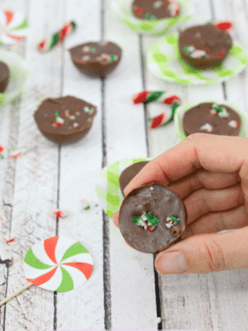 Hand holding small portion of chocolate fudge with candy canes on top with more fudhe on green cupcake holders behind it