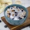 Healthy protein oatmeal in a blue bowl with blueberries and pecans sitting on a cutting board with a yellow napkin and protein powder behind it