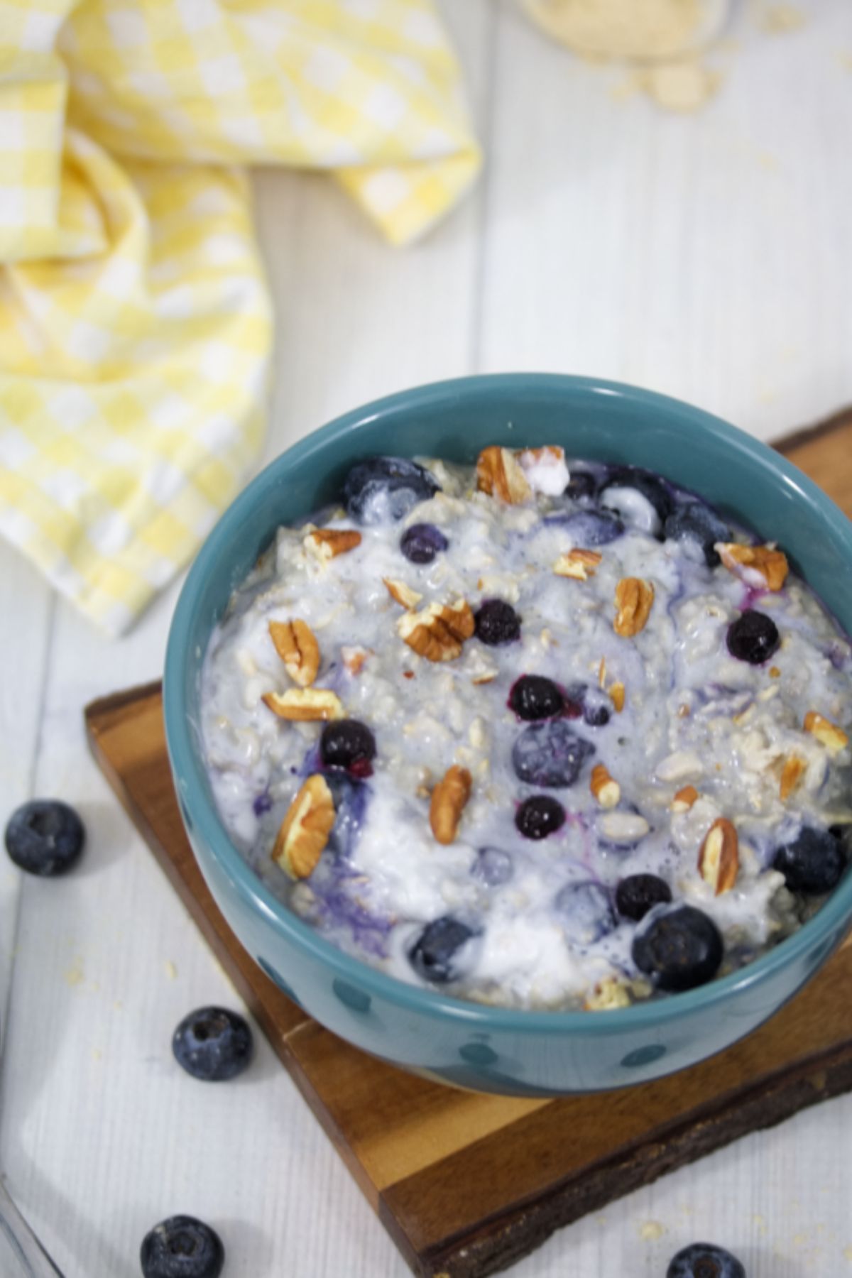 Protein oatmeal in a blue bowl with blueberries and pecans sitting on a cutting board with a yellow napkin and protein powder behind it.