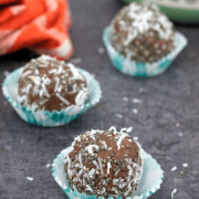 Featured image of three chocolate protein balls in blue cupcake liners with more on an orange linen behind them and a tub of protein powder
