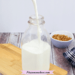Glass milk jug on light wood cutting board with homemade non-dairy milk being poured in and a purple bowl of granola behind it