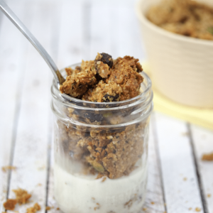 Homemade granola in a small mason jar with milk in it and a spoon in the top. More granola scattered around the jar and in a beige bowl behind it