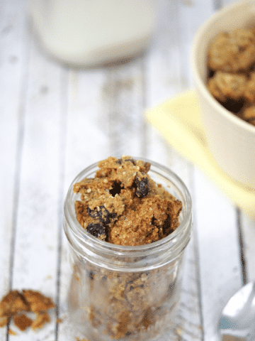 Mason jar with homemade protein granola ith more granola in a tan bowl on a yellow napkin behind it and a milk jug behind that