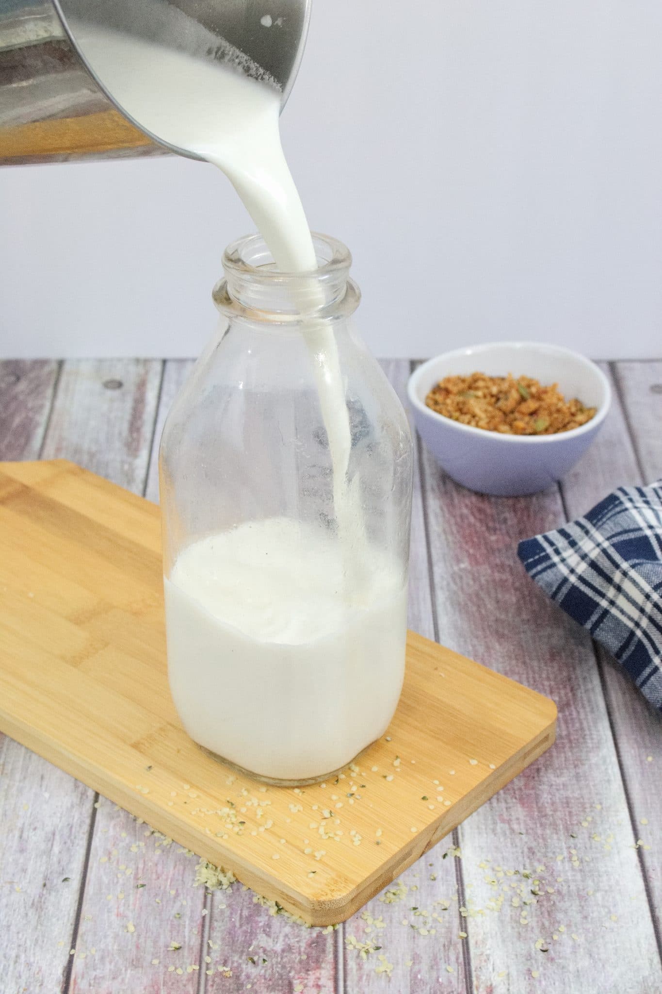Glass milk jug on light wood cutting board with homemade non-dairy milk being poured in and a purple bowl of granola behind it
