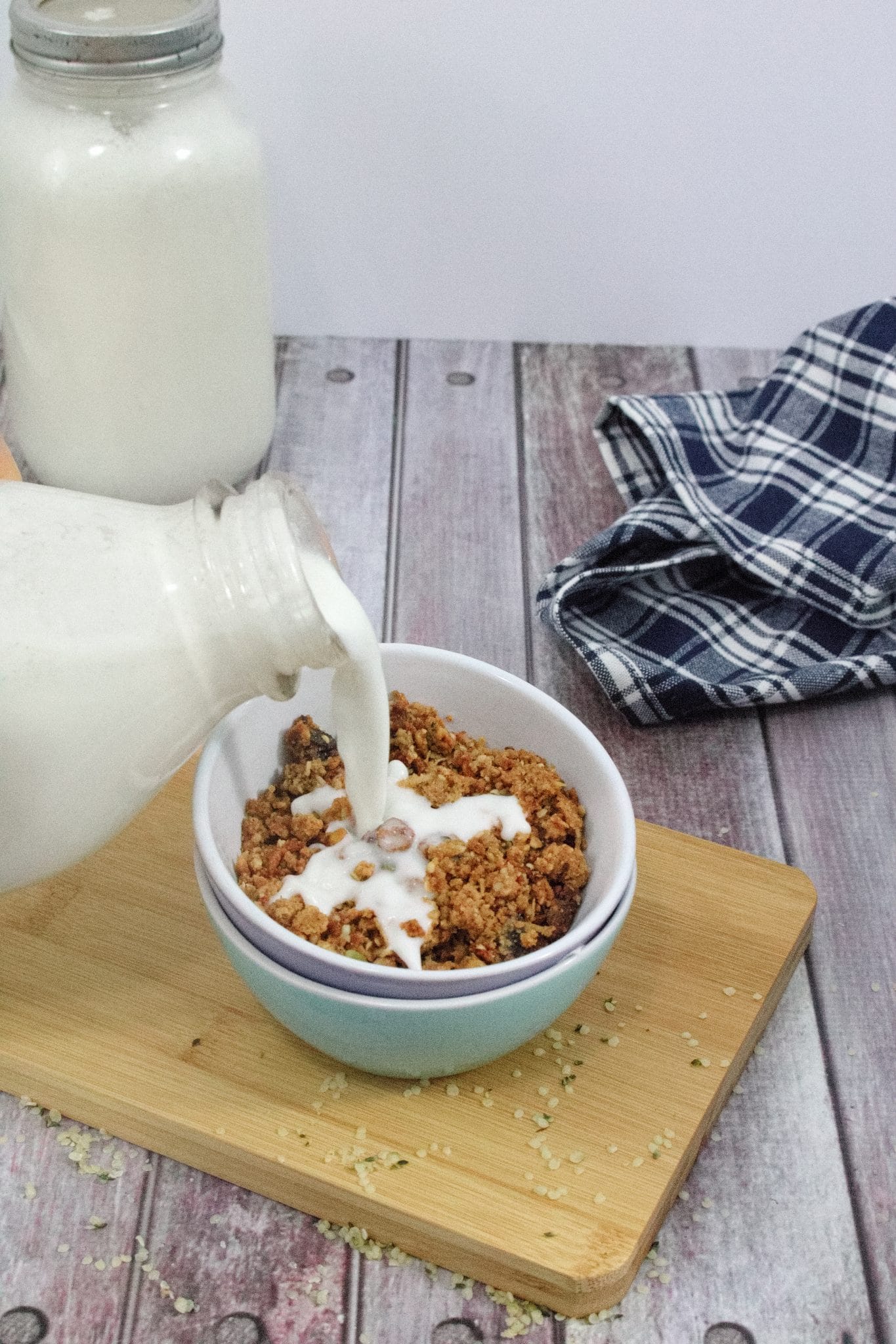 Double stacked bowls on a cutting board with granola in them and homemad hemo milk being poured out of a glass jug on top