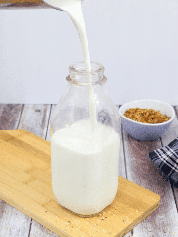 Glass milk jug on light wood cutting board with homemade non-dairy milk being poured in and a purple bowl of granola behind it