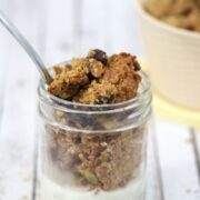 Homemade granola in a small mason jar with milk in it and a spoon in the top. More granola scattered around the jar and in a beige bowl behind it.