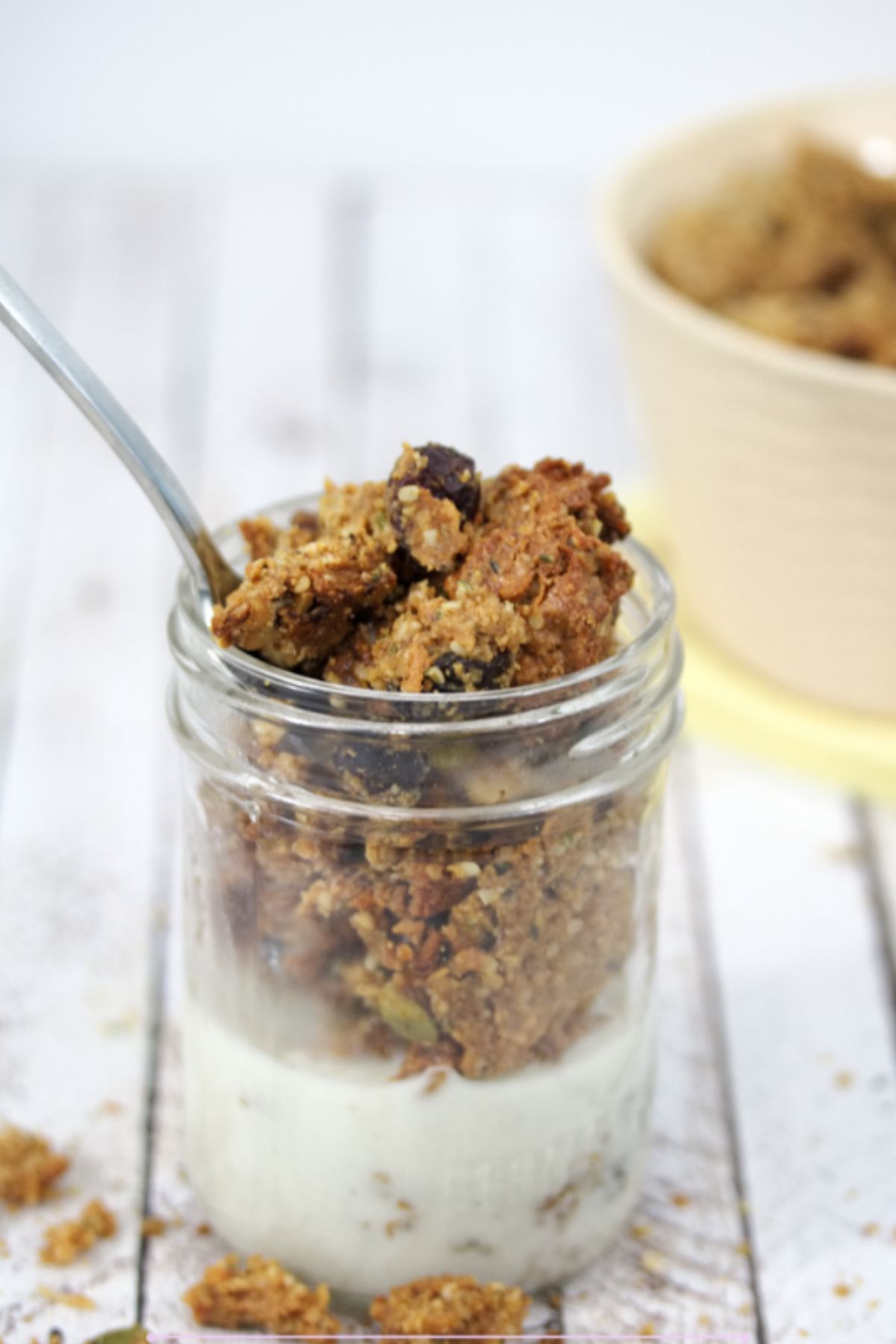 Homemade granola in a small mason jar with milk in it and a spoon in the top. More granola scattered around the jar and in a beige bowl behind it.