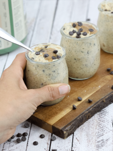 healthy cookie dough overnight oats with nut butter and chocolate chips in three mini mason jars with a hand grabbing one jar