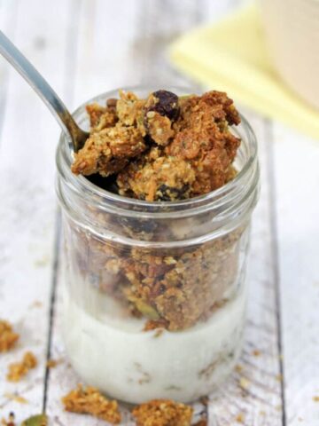 Homemade granola in a small mason jar with milk in it and a spoon in the top. More granola scattered around the jar and in a beige bowl behind it
