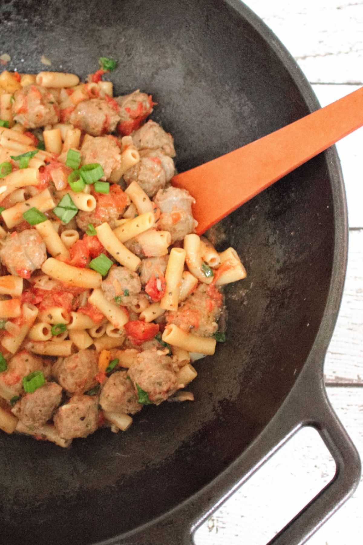 Cast iron pan with tomato and sausage pasta on gluten-free noodles topped with green onions and spinach with a wooden spoon in the pot.