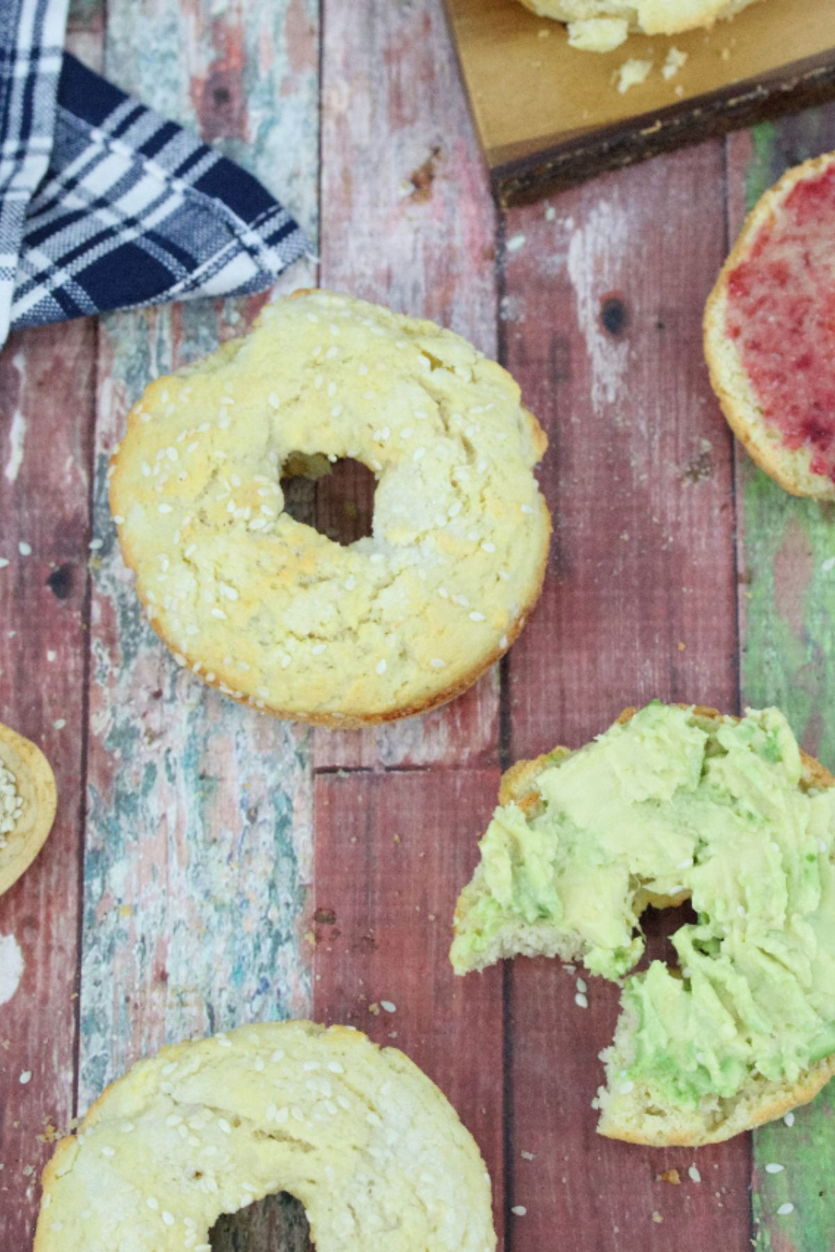 Multiple protein bagels, some sliced with avocado or jam on them with a blue linen and cutting board behind them.