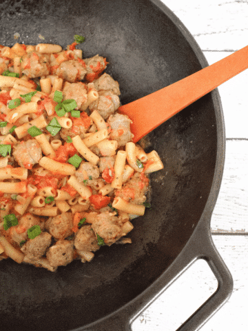 Cast iron wok with tomato and sausage pasta on gluten-free noodles topped with spinach and a wooden spoon in it.