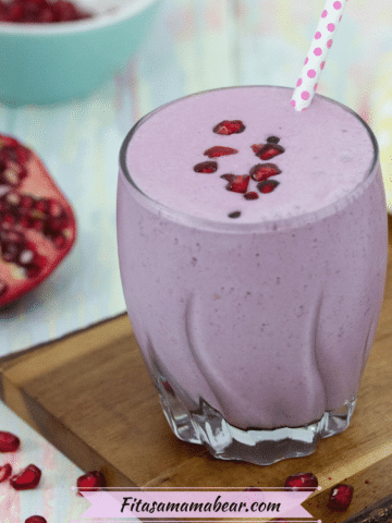 Purple pomegranate smoothie in small glass topped with polkadot straw and pomegranate seeds on a cutting board with a sliced pomegranate behind it