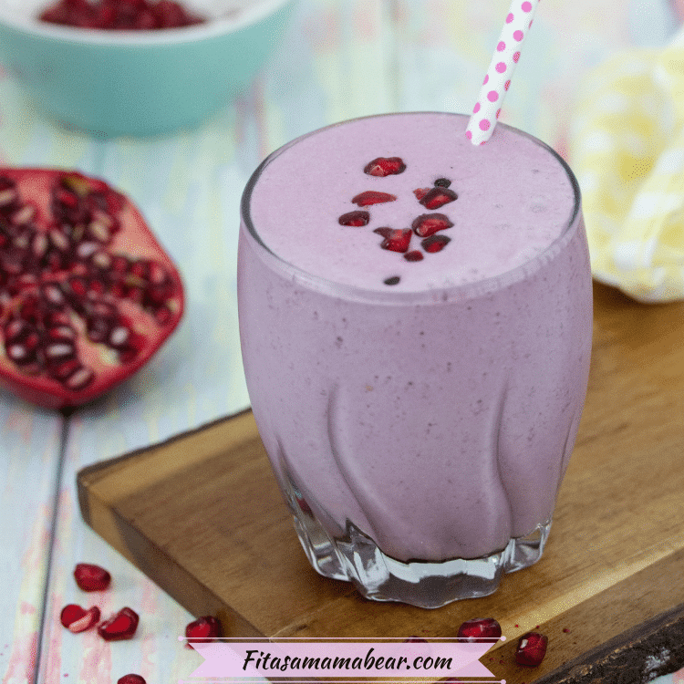 Purple pomegranate smoothie in small glass topped with polkadot straw and pomegranate seeds on a cutting board with a sliced pomegranate behind it