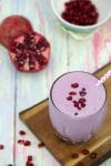 Purple pomegranate smoothie in small glass topped with polkadot straw and pomegranate seeds on a cutting board with a sliced pomegranate behind it
