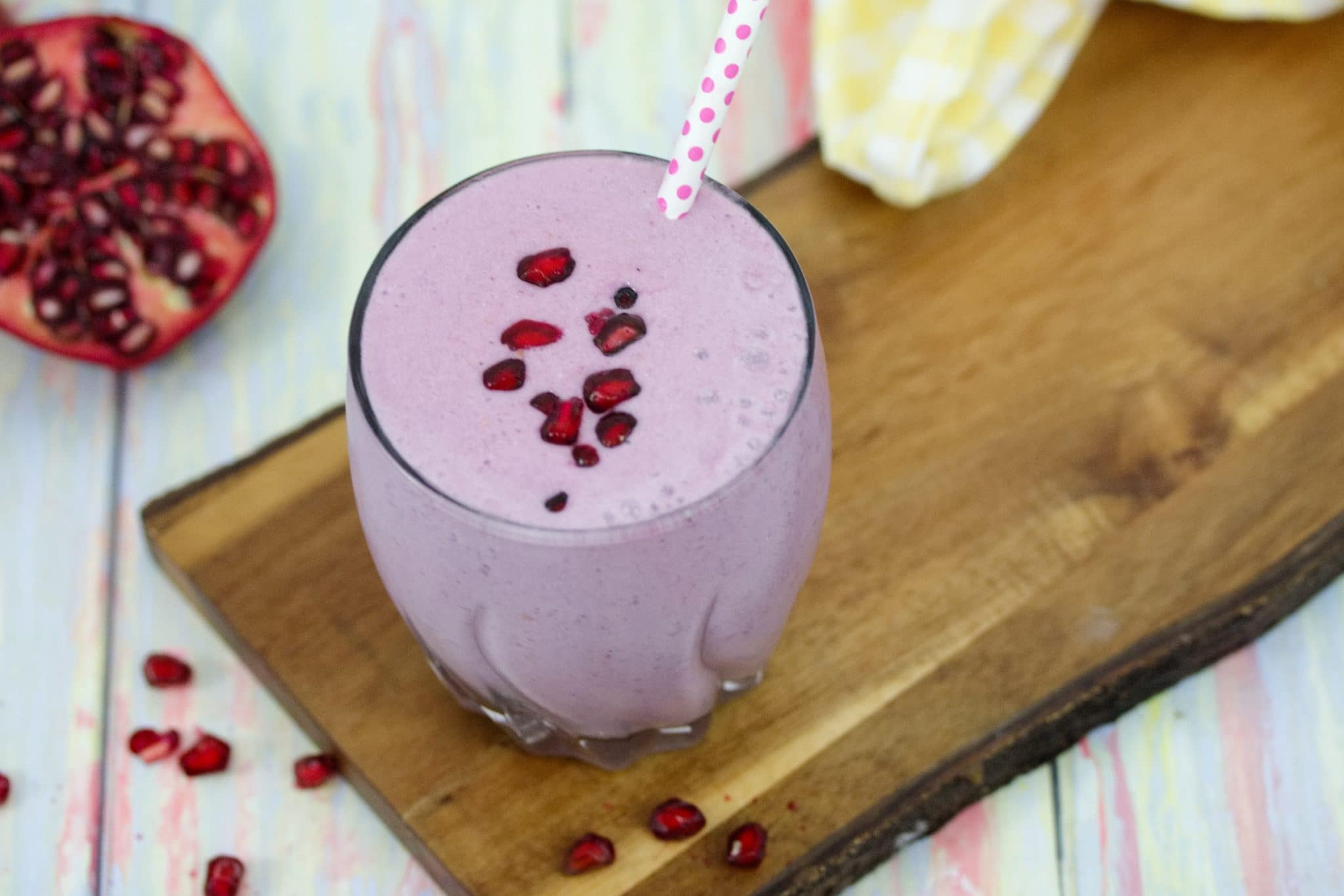 Purple pomegranate smoothie in small glass topped with polkadot straw and pomegranate seeds on a cutting board with a sliced pomegranate behind it