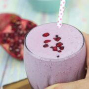 Pomegranate smoothie in a glass topped with arils and a polka dot straw with a hand holding the glass on a cutting board.