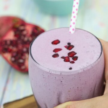 Pomegranate smoothie in a glass topped with arils and a polka dot straw with a hand holding the glass on a cutting board.