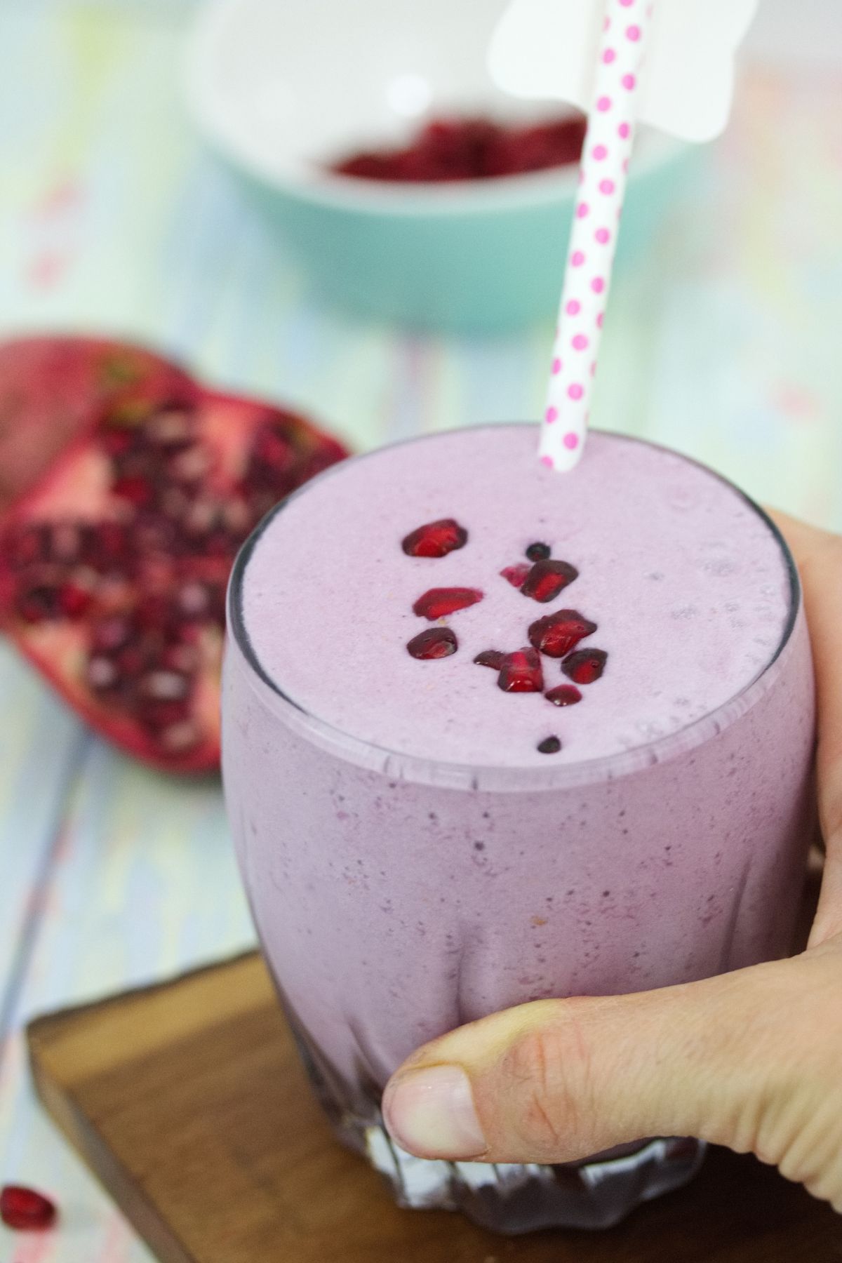 Pomegranate smoothie in a glass topped with arils and a polka dot straw with a hand holding the glass on a cutting board.