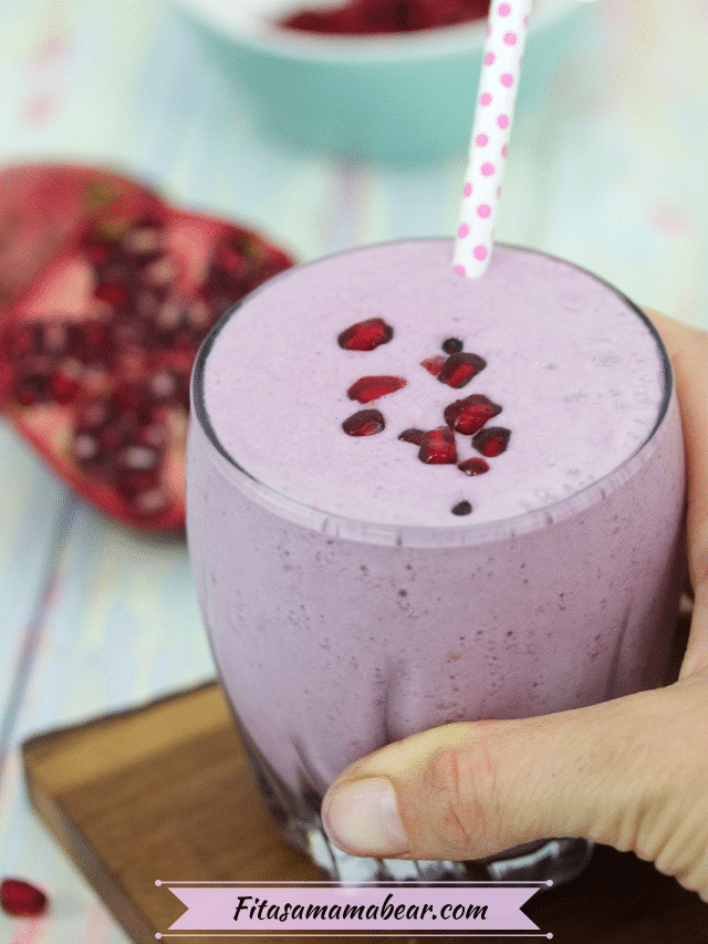 Pomegranate smoothie in a glass topped with arils and a polka dot straw with a hand holding the glass on a cutting board