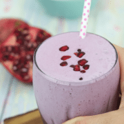 Pomegranate smoothie in a glass topped with arils and a polka dot straw with a hand holding the glass on a cutting board