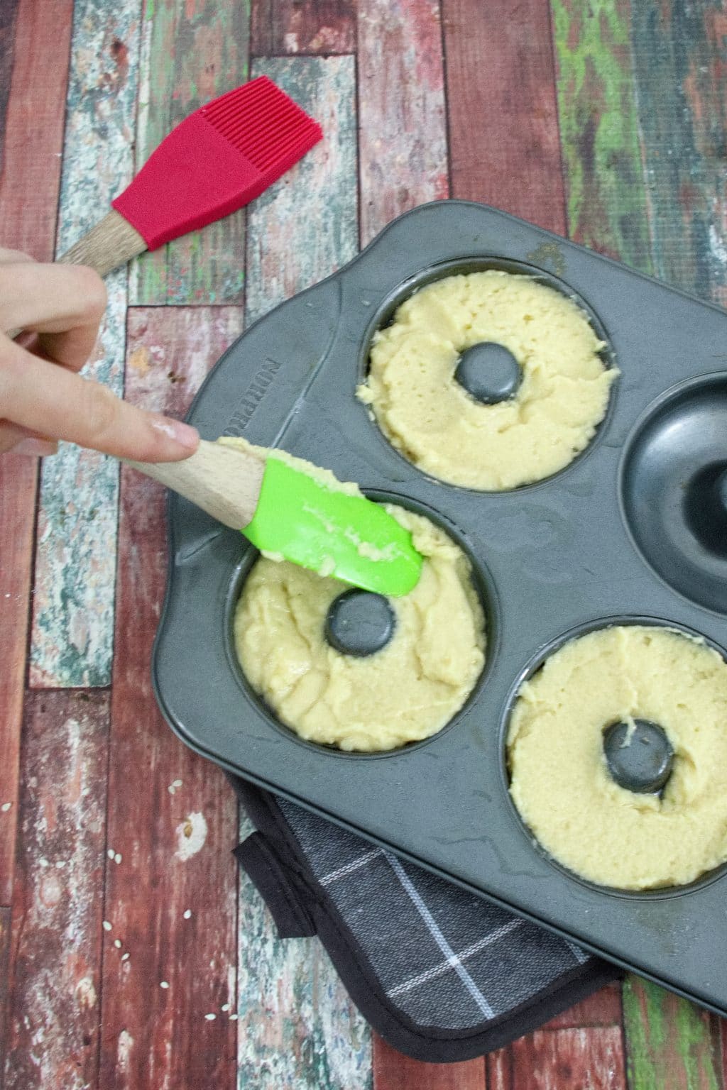 Donut tray with homemade bagels in it and a green silicone spatula pressing the bagels down