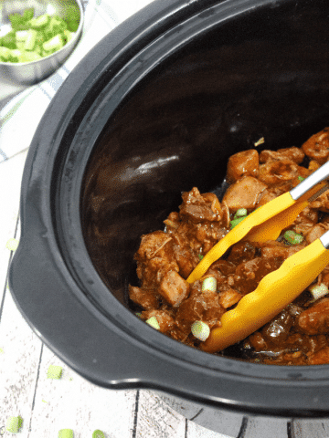 Black crock pot with gluten-free teriyaki chicken and yellow tongs scooping some out topped with green onions