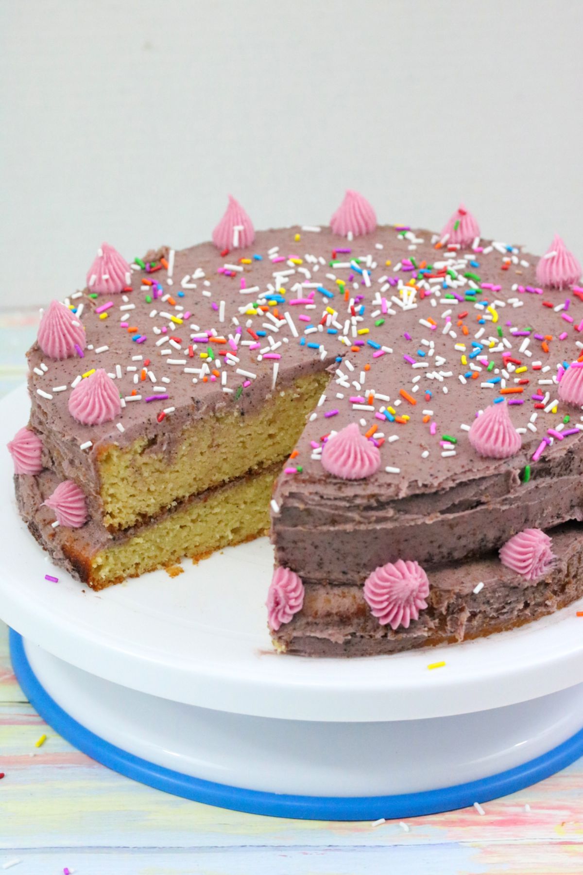 Dairy-free birthday cake with purple icing and pink icing flowers and covered in sprinkles on a cake turntable.