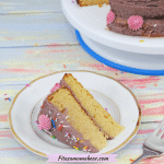 A slice of vanilla birthday cake with purple icing and sprinkles on a white plate with a fork beside it and more cake on a turntable behind it