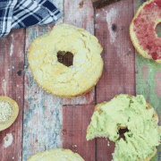 Multiple, gluten-free protein bagels, some sliced with avocado or jam on them with a blue linen and cutting board behind them