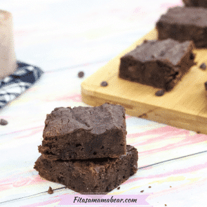 Two chocolate brownies stacked with more brownies on a cutting board and a scoop of protein powder on a blue linen