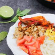 Chicken fajita bowl on rice with tomatoes and peppers in a white bowl with lime on the side.
