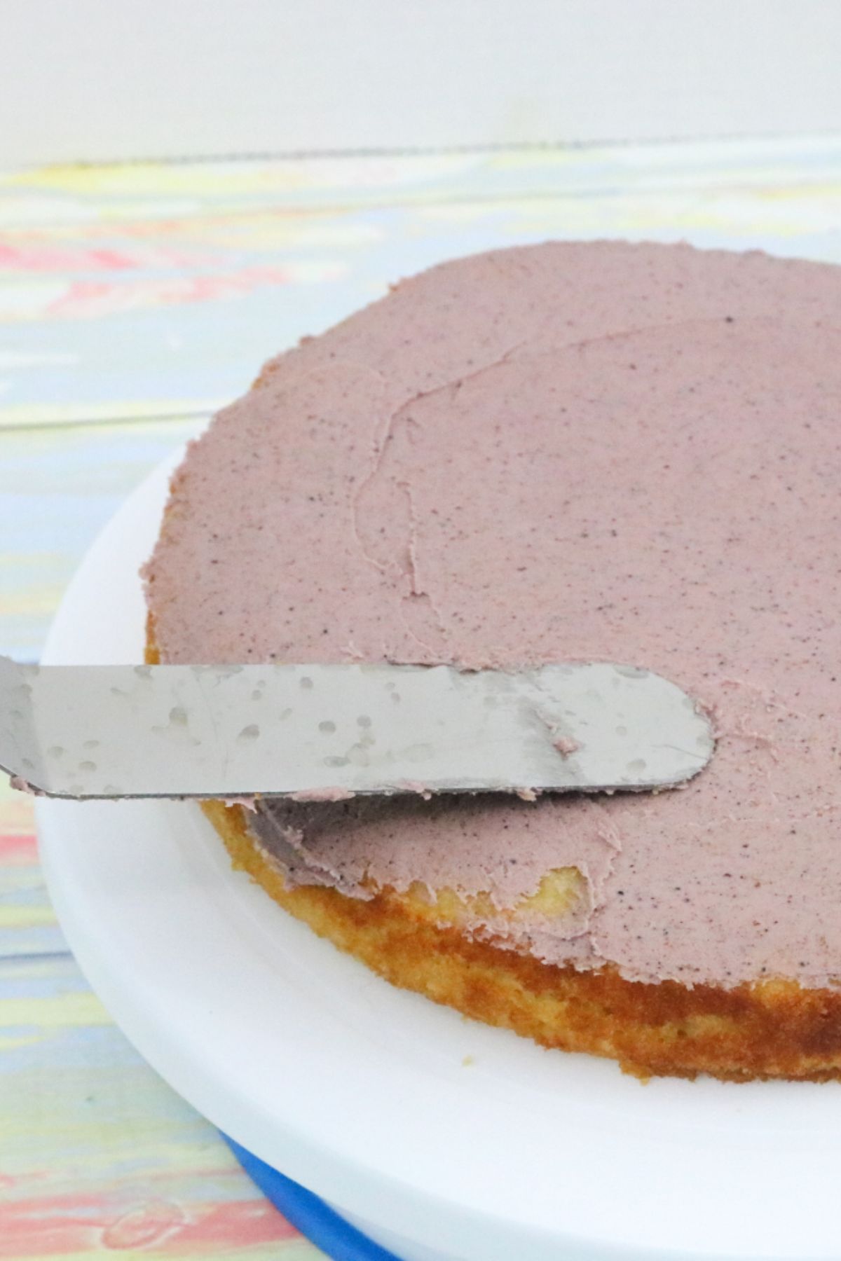 Small dairy-free cake on a turntable being iced purple with an icing spatula.