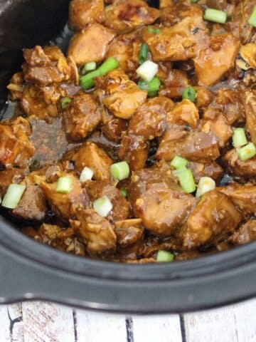 Black slow cooker with teriyaki chicken being scooped by yellow tongs and topped with green onions