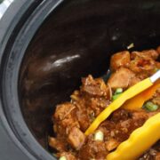 Black slow cooker with teriyaki chicken being scooped by yellow tongs and topped with green onions
